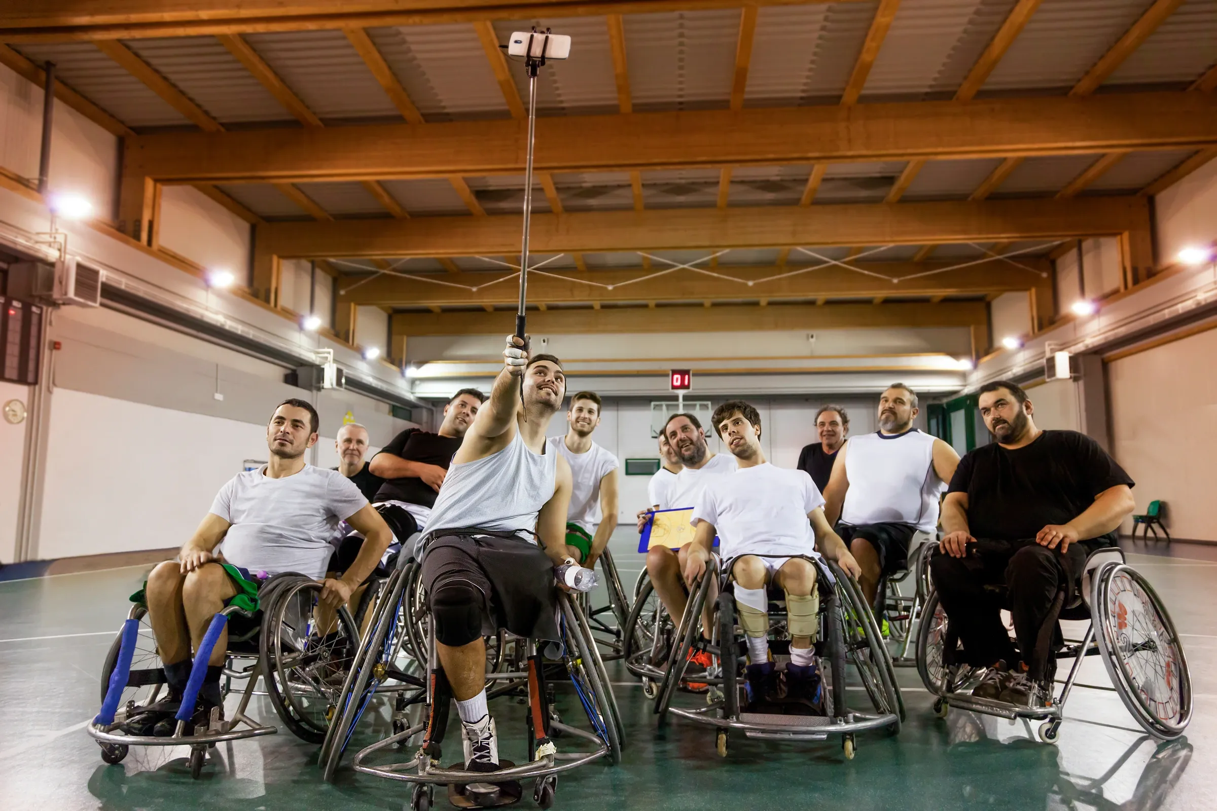 Eine Gruppe von Rollstuhlbasketballern macht gemeinsam ein Selfie mit einem Selfiestick in der Sporthalle.