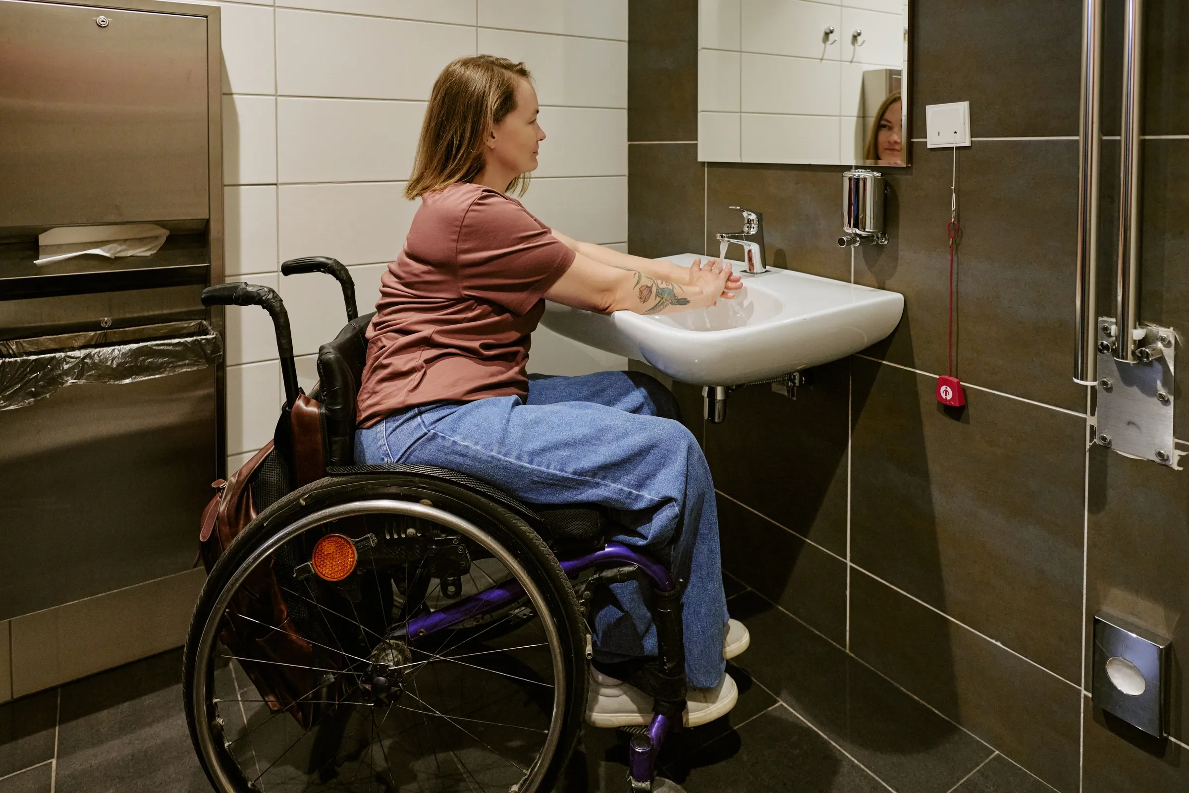 Eine Frau im Rollstuhl wäscht sich die Hände an einem unterfahrbaren Waschbecken in einer barrierefreien Toilette.